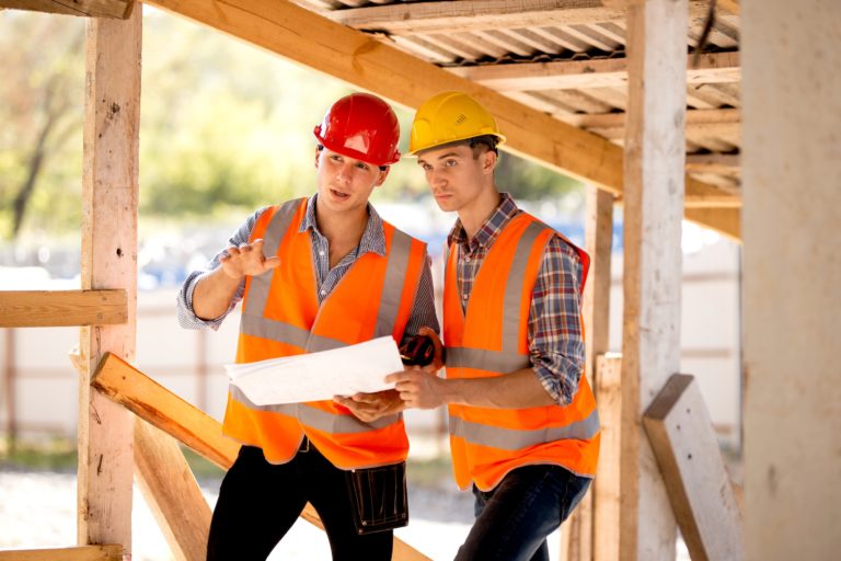 Två personer i orange arbetsvästar står vid byggarbetsplats och tittar på ritning